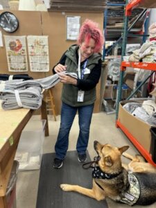 Kelly Ann sorts towels at Bridging while her service dog Dino lays attentively on the floor beside her.